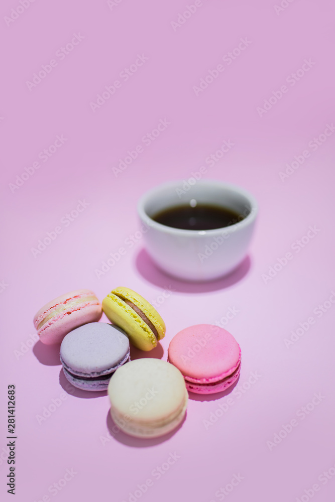 Overview of sweet colorful macaroon biscuits lying on pastel pink background. A mug with black coffee beside.