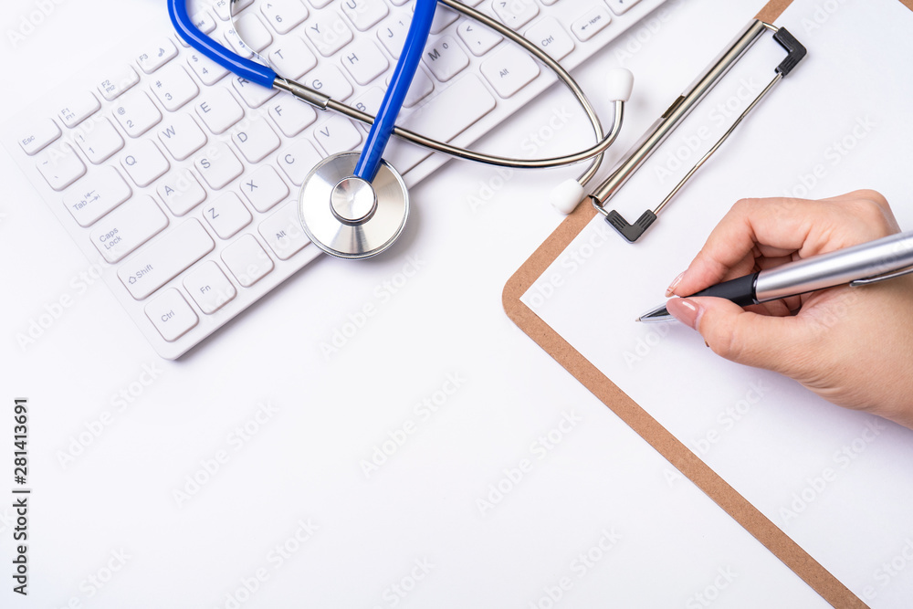 Female doctor writing a medical record case over clipboard on white working table with stethoscope, computer keyboard. Top view, flat lay, copy space Female doctor writing a medical record case over clipboard on white working table with stethoscope, computer keyboard. Top view, flat lay, copy space