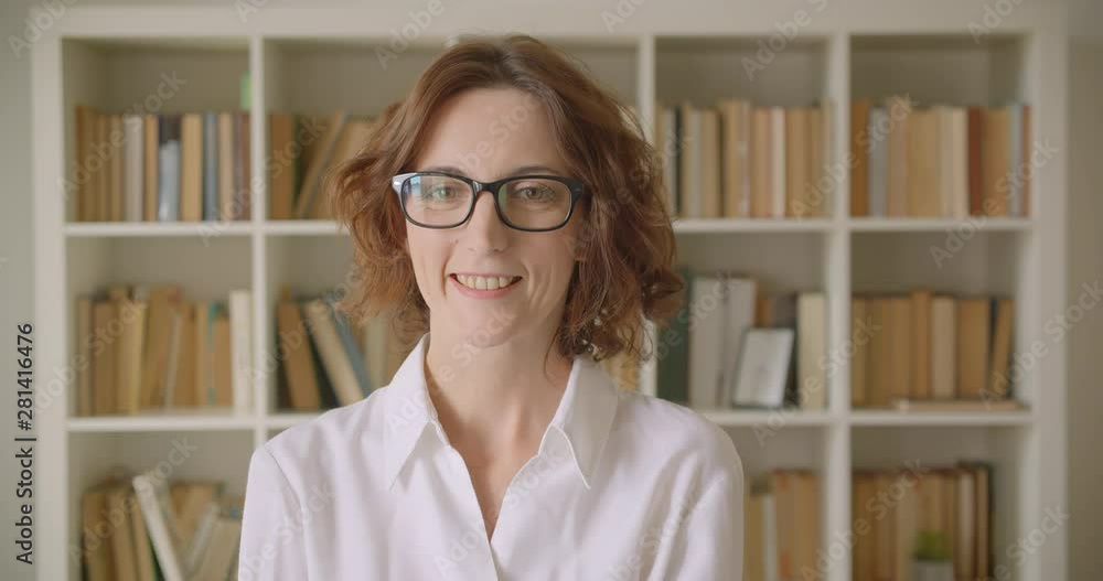 Closeup portrait of adult redhead attractive caucasian businesswoman looking at camera smiling happily indoors with bookshelves on the background Closeup portrait of adult redhead attractive caucasian businesswoman looking at camera smiling happily indoors with bookshelves on the background