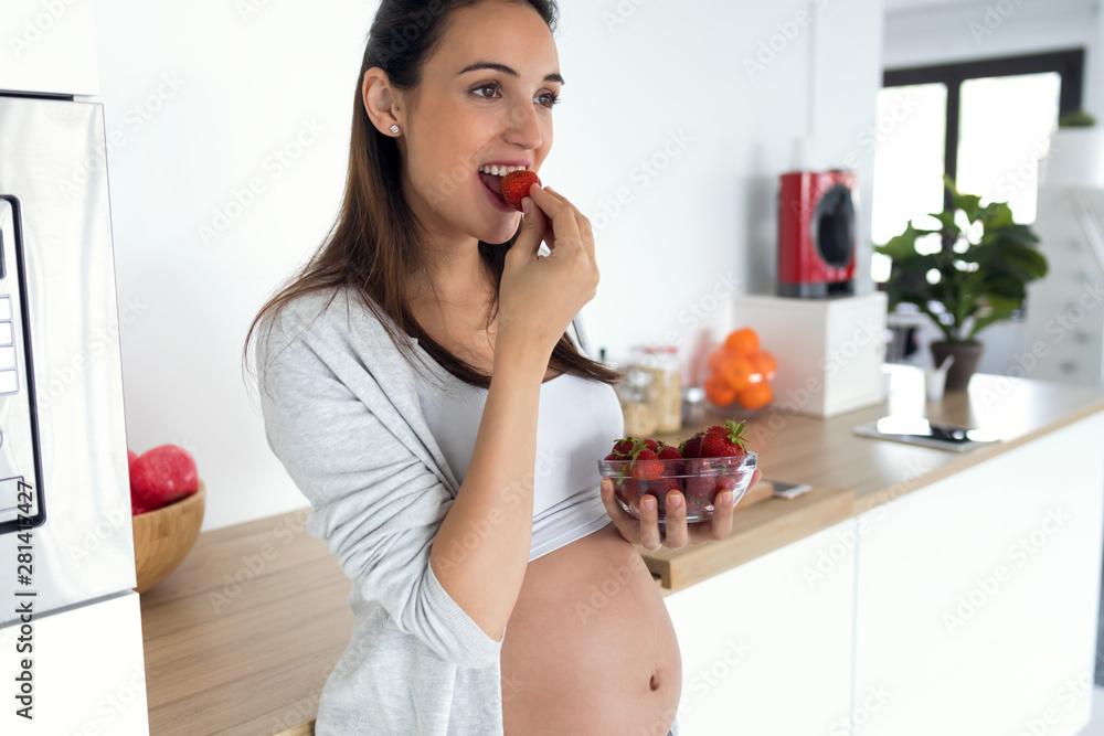 Beautiful pregnant young woman eating strawberries out of bowl in the kitchen at home. Beautiful pregnant young woman eating strawberries out of bowl in the kitchen at home.