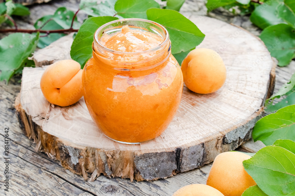 Apricot jam in a glass jar next to a ripe apricot and green leaves on a wooden surface.