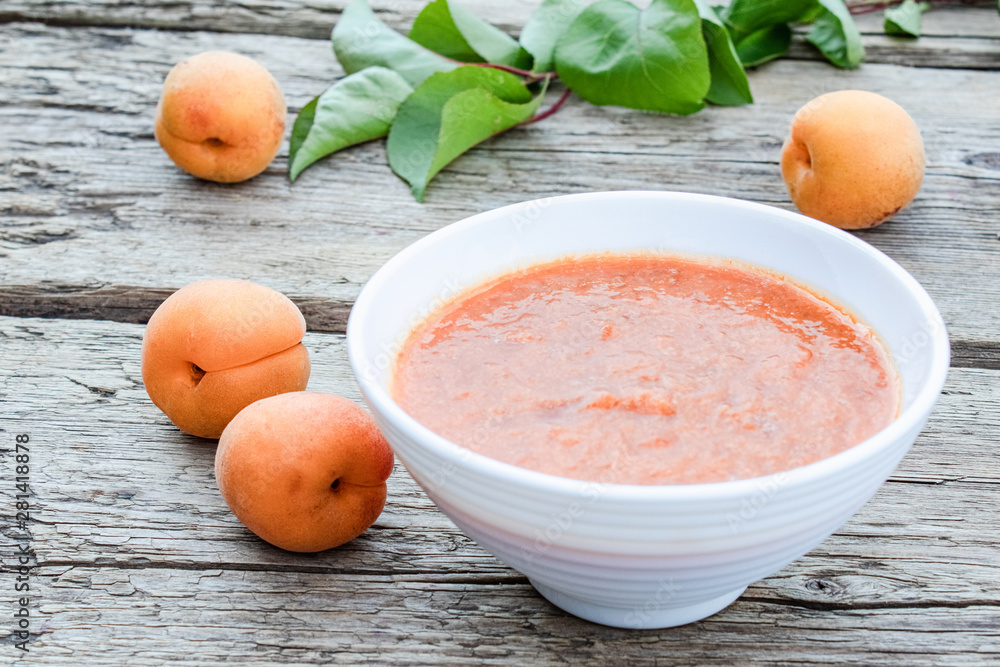 White bowl with homemade apricot jam on a wooden surface near ripe apricot and green leaves. White bowl with homemade apricot jam on a wooden surface near ripe apricot and green leaves.