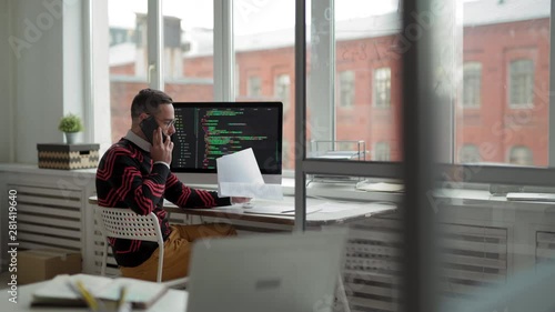 Tracking left of middle-aged Caucasian businessman sitting at desktop at computer and having phone call concerning papers in his hands