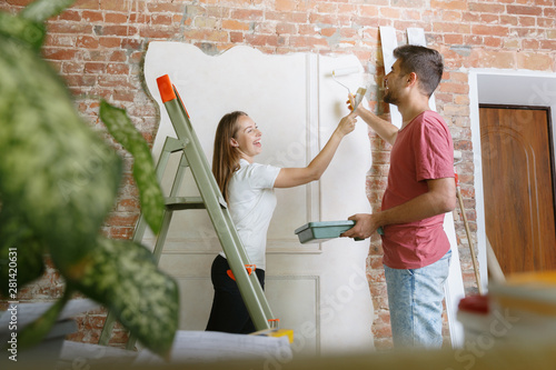 Young couple doing apartment repair together themselves. Married man and woman doing home makeover or renovation. Concept of relations, family, love. Painting the wall together and laughting.
