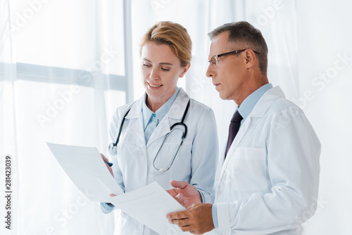 cheerful doctor holding paper near handsome coworker in glasses