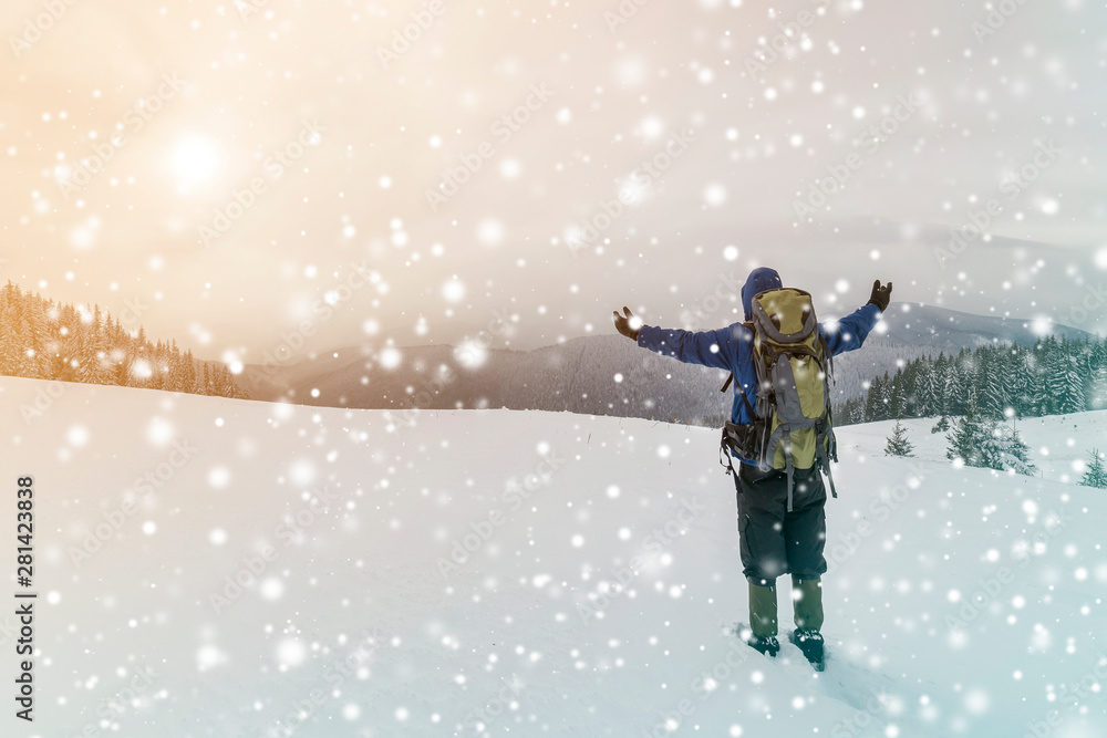 Back view of tourist hiker in warm clothing with backpack standing with raised arms on clearing covered with snow on spruce forest mountain and cloudy sky copy space background. Back view of tourist hiker in warm clothing with backpack standing with raised arms on clearing covered with snow on spruce forest mountain and cloudy sky copy space background.
