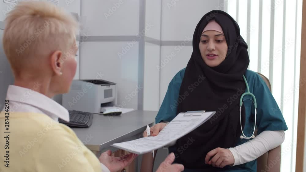 Lockdown of young Muslim doctor holding form in her hands and showing with pen to middle-aged Caucasian woman how to fill it in and then giving form to patient Lockdown of young Muslim doctor holding form in her hands and showing with pen to middle-aged Caucasian woman how to fill it in and then giving form to patient