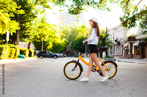 Girl teenager in a hat with a yellow bike walks through the summer city.