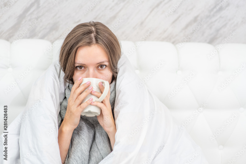 Beautiful sick woman sitting on the bed wrapped in a white blanket and drink hot tea. Empty space for text Beautiful sick woman sitting on the bed wrapped in a white blanket and drink hot tea. Empty space for text