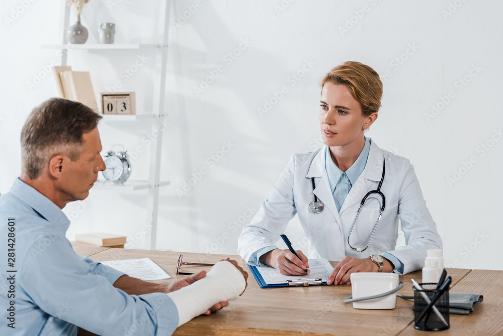 Obraz premium attractive doctor looking at patient with broken arm while writing diagnosis in clinic