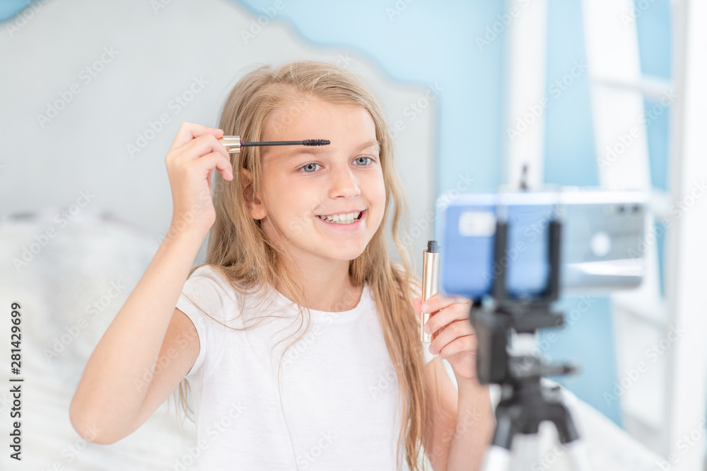 Smiling girl applies mascara on eyelashes and record video for her followers. Kids blogger concept