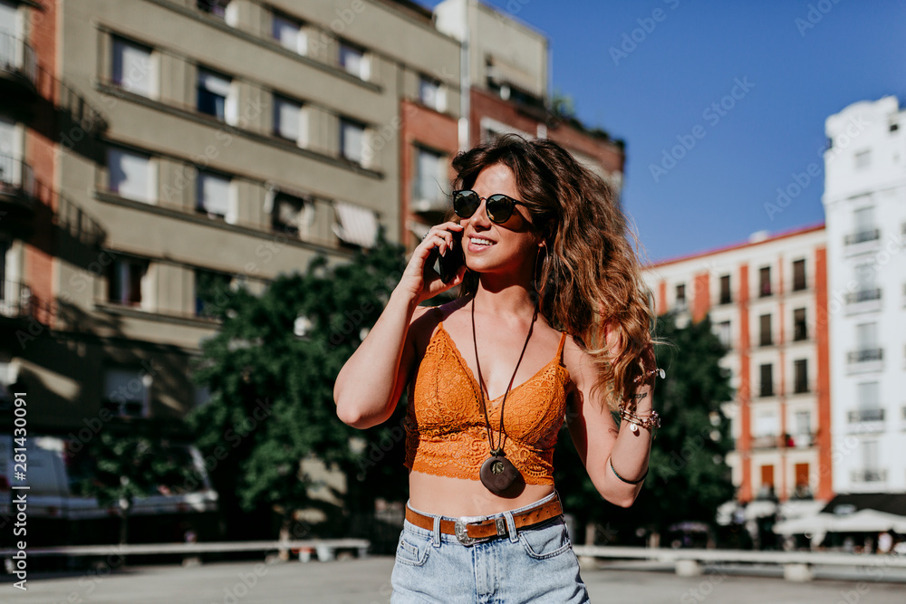 Beautiful young caucasian woman walking at the city street on a sunny day. Talking on mobile phone. Happy face smiling. Urban lifestyle