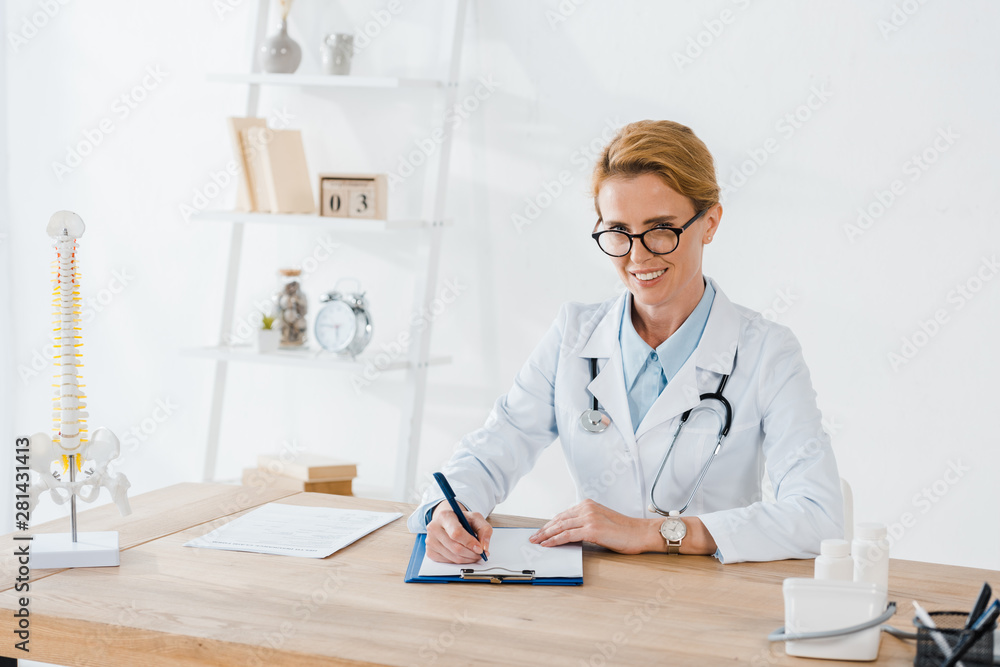 happy doctor in glasses writing diagnosis near spine model in clinic happy doctor in glasses writing diagnosis near spine model in clinic