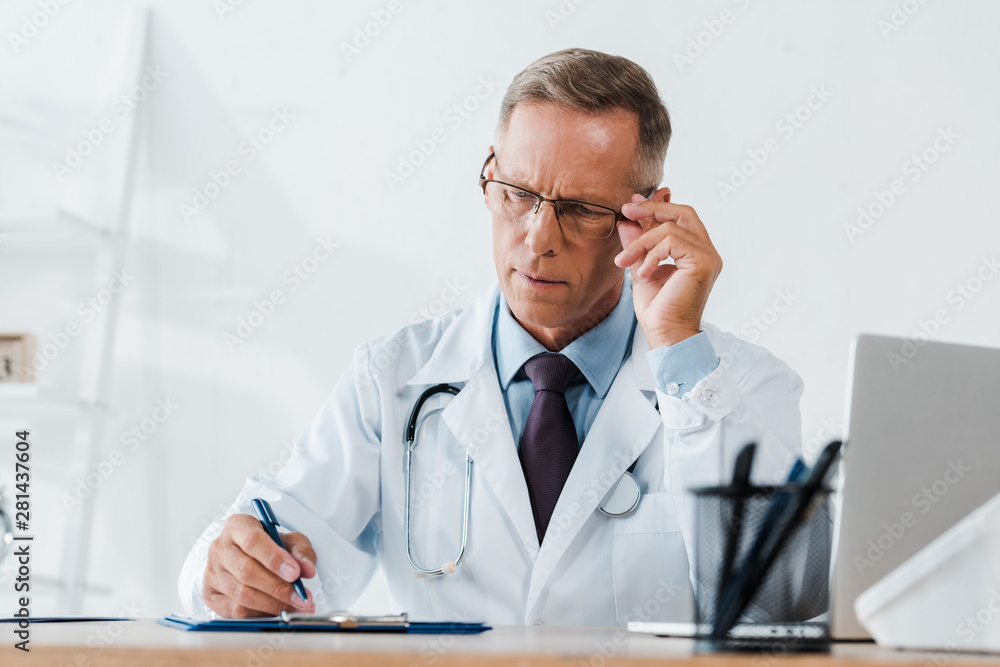 selective focus of handsome doctor touching glasses while writing on clipboard in hospital