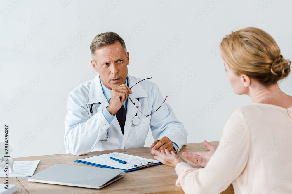 selective focus of doctor in white coat looking at woman and holding glasses in hospital selective focus of doctor in white coat looking at woman and holding glasses in hospital