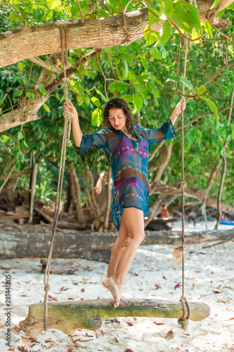 slim sexy girl model in a swimsuit posing on a wooden swing tied to a tree. On the background of the beach of a tropical island
