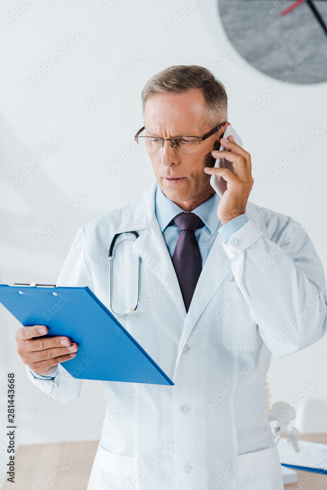 serious doctor in glasses looking at clipboard and talking on smartphone serious doctor in glasses looking at clipboard and talking on smartphone