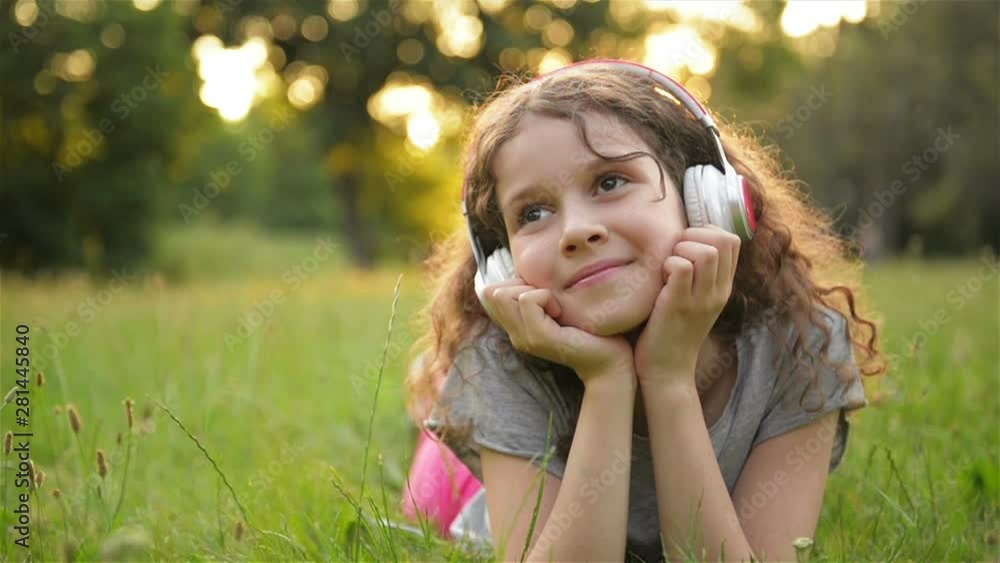Portrait of Beautiful Caucasian Little Girl Spending Time In Public Park. Student Kid Having a Lot of Fun Relaxing Outdoors and Listening To Music With Headphones. Portrait of Beautiful Caucasian Little Girl Spending Time In Public Park. Student Kid Having a Lot of Fun Relaxing Outdoors and Listening To Music With Headphones.