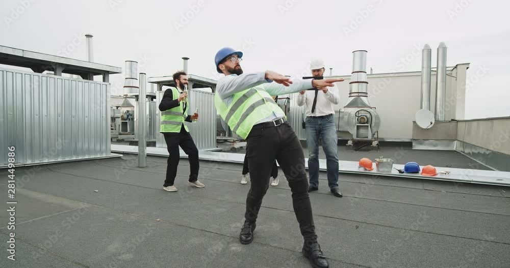 Three construction people , a handsome man and a good looking woman dance slowly behind a guy who is dancing professionally , while getting filmed on top of an architecture site Three construction people , a handsome man and a good looking woman dance slowly behind a guy who is dancing professionally , while getting filmed on top of an architecture site
