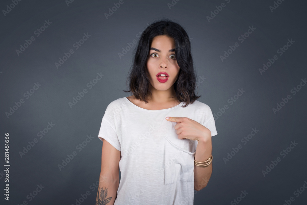 Surprised beautiful female student being in stupor shocked, has astonished expression pointing at herself with finger saying: Who me? Isolated over gray background. Surprised beautiful female student being in stupor shocked, has astonished expression pointing at herself with finger saying: Who me? Isolated over gray background.