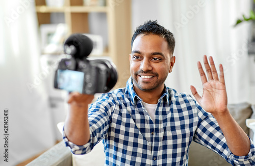 blogging, videoblog and people concept - smiling indian male video blogger with camera videoblogging and waving hand at home