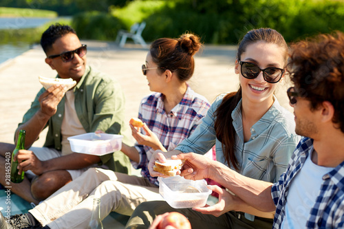 leisure and people concept - group of happy friends having picnic and eating sandwiches on lake pier in summer