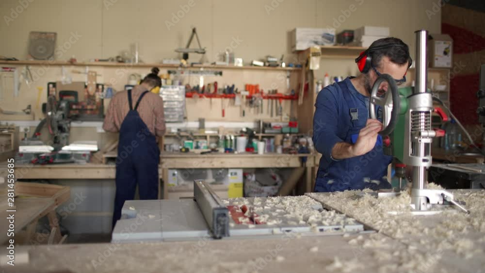 Tracking right of young carpenter on background doing something with his hands, while his middle-aged colleague on foreground working with drill Tracking right of young carpenter on background doing something with his hands, while his middle-aged colleague on foreground working with drill