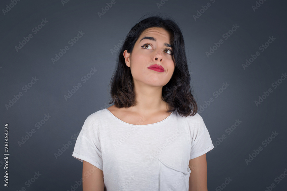 Troublesome puzzled Caucasian woman frowns face, bites lip, raises eyebrows, looks up, has problems, dressed in fashionable clothes, isolated over gray background, has regret look. Troublesome puzzled Caucasian woman frowns face, bites lip, raises eyebrows, looks up, has problems, dressed in fashionable clothes, isolated over gray background, has regret look.