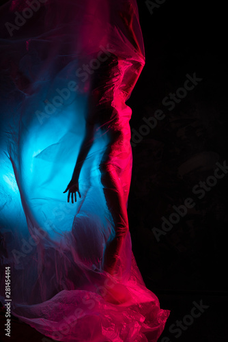 Fotografiet Slim girl dressed in underwear emotionally posing, wrapped in fluttering in the wind and fitting her graceful young body cellophane film, in blue and red light