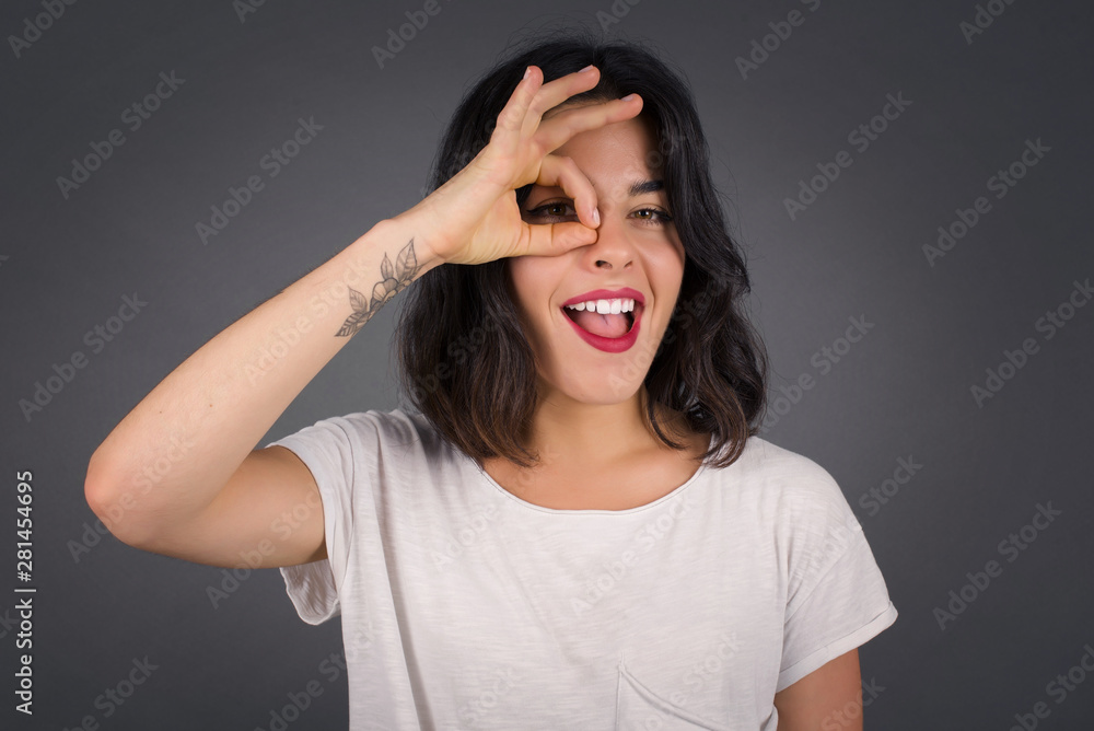 Beautiful young blonde woman with happy face smiling doing ok sign with hand on eye looking through finger. Wearing casual clothes and standing. Beautiful young blonde woman with happy face smiling doing ok sign with hand on eye looking through finger. Wearing casual clothes and standing.