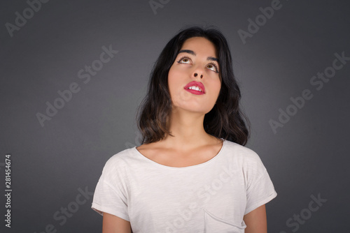 portrait of mysterious charming middle aged brunette female with straight hair looking up with enigmatic smile. Beautiful smiling girl looking up standing against gray wall.