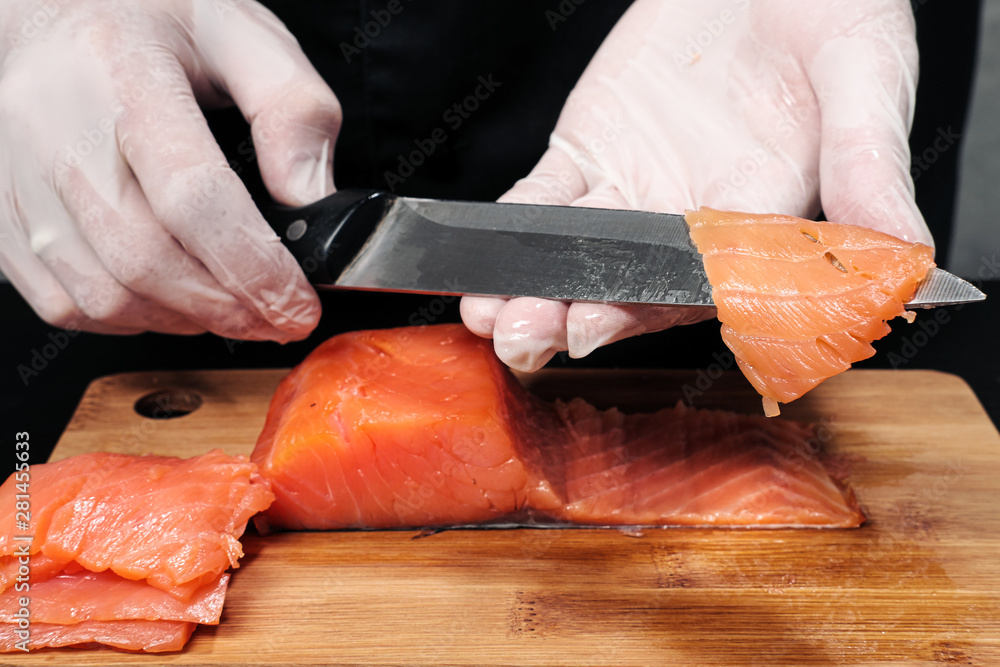 Hands cooks close-up. The chef cuts with a knife a red fish, smoked salmon on a wooden cutting board.