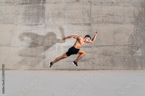 Muscular shirtless caucasian male athlete does an explosive starting sprint against a grungy concrete wall background 