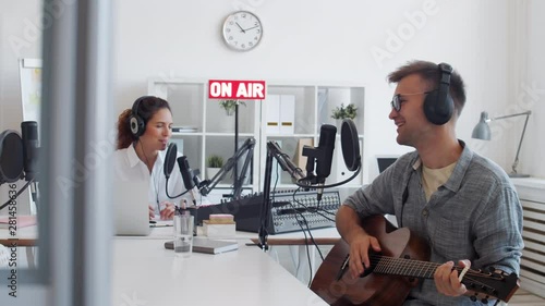 Medium shot of male guest musician sitting at microphone, finishing playing guitar and singing and then female Caucasian radio host sitting on background starting speaking into mike photo