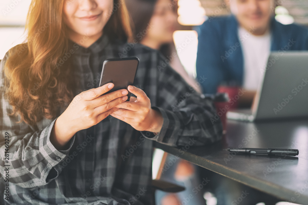 A beautiful asian woman using and looking at mobile phone with colleaques working on laptop in background A beautiful asian woman using and looking at mobile phone with colleaques working on laptop in background