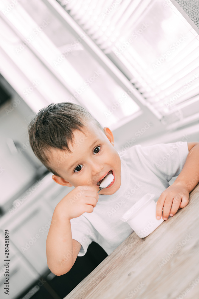 Cute boy in the kitchen eating yogurt from a white yogurt container, a place for advertising Cute boy in the kitchen eating yogurt from a white yogurt container, a place for advertising