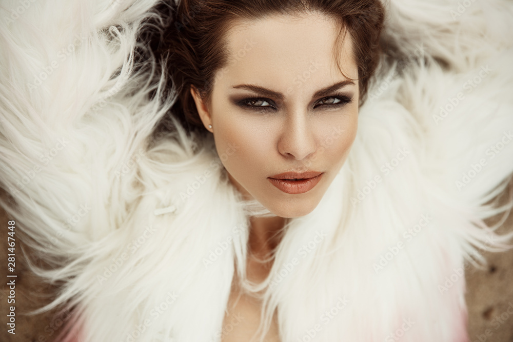 Fashionable and stylish portrait of a beautiful and magnificent young brunette woman with sexy makeup (full lips, smokey eyes) and loose hair in a white gorgeous fur coat