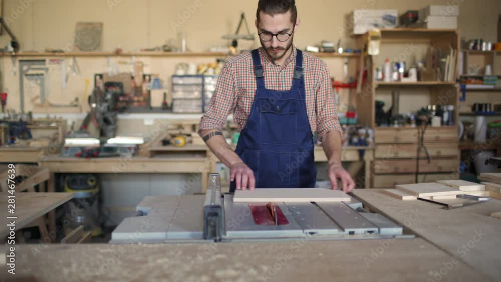 Tracking right of young joiner sawing with saw lump of wood, while his colleague on background walking by with papers in his hands Tracking right of young joiner sawing with saw lump of wood, while his colleague on background walking by with papers in his hands