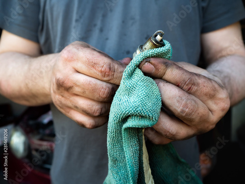 a man in dirty clothes and with grubby hands, holding a rag and a spark plug, wiping the car parts