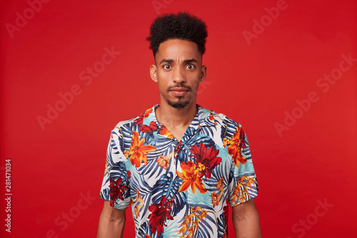 Young wondered African American man, wears in Hawaiian shirt, looks at the camera with surprised expression and wide opened eyes, stands over red background.