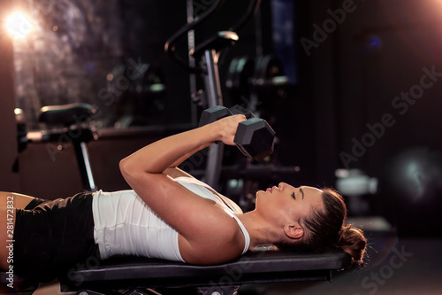 Strong young sexy girl lying on bench with weights on hands in gym