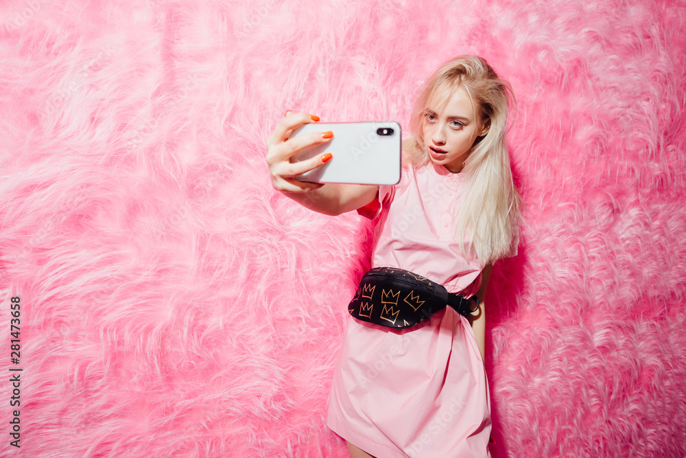Bright young girl blogger dressed in fashion pink dress takes a selfie on  her smartphone on the background of pink fur wall in the show room Stock  Photo | Adobe Stock