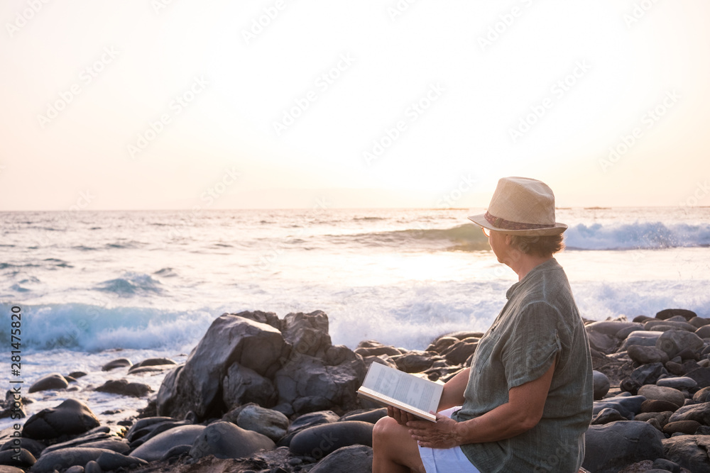 Senior woman alone sitting on the beach of pebbles reading a book. 70 years old. Waves of the ocean. Sunset light