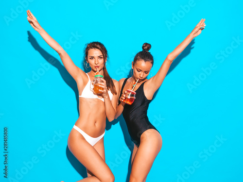 Two beautiful sexy smiling hipster women in summer white and black swimwear bathing suits.Trendy girls going crazy.Funny models isolated on blue.Drinking fresh cocktail smoozy drink.Raising hands