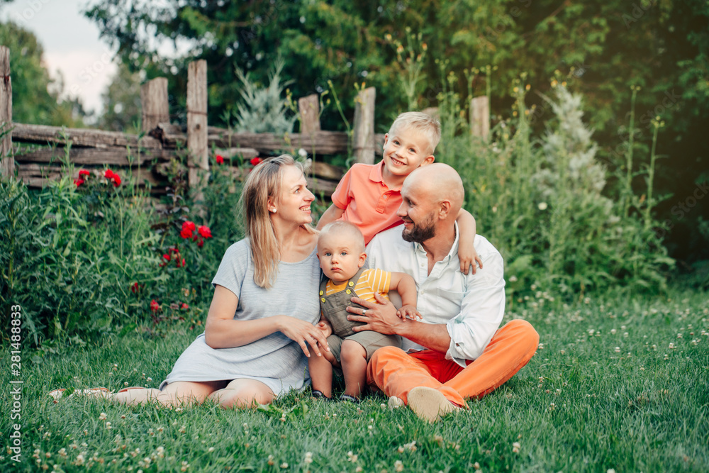Fototapeta premium Beautiful happy Caucasian Ukrainian family of four people in park on summer day. Mother and father hugging their two sons brothers siblings. Lifestyle authentic natural emotions moment.