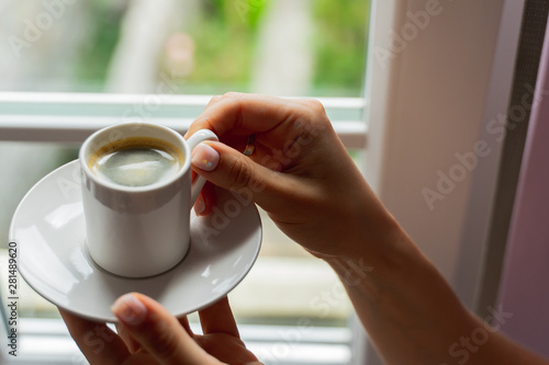 woman hands holding cup of coffee near window in morning sunlight.