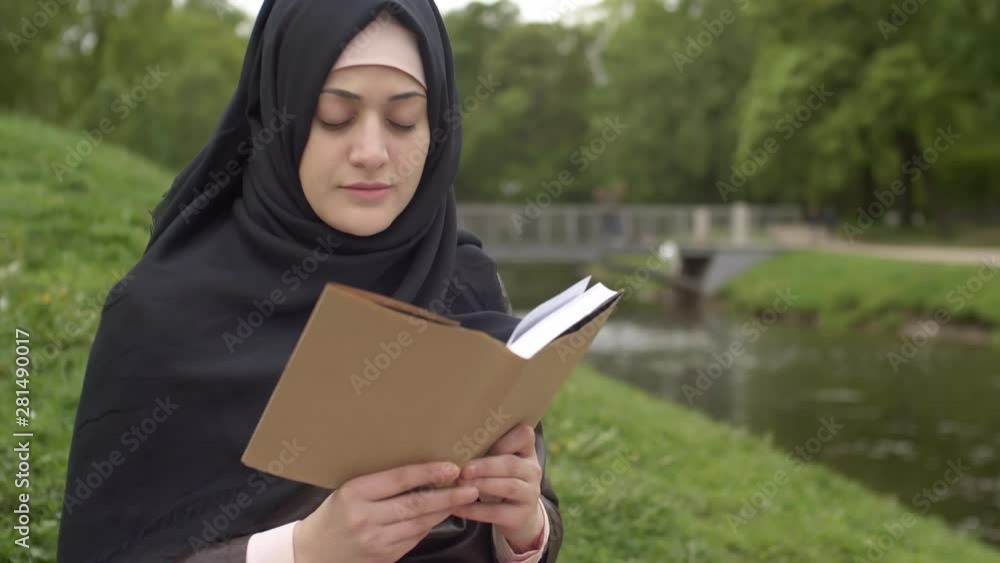 Panning of young Muslim woman wearing veil sitting alone on lawn in park and reading book Panning of young Muslim woman wearing veil sitting alone on lawn in park and reading book