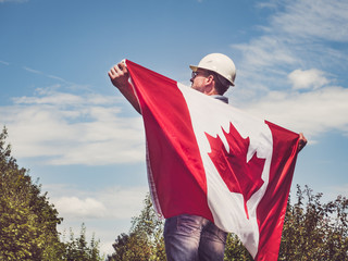 Handsome engineer, holding Canadian Flag in the park against the backdrop of green trees and the setting sun, looking into the distance. Close-up. Concept of labor and employment