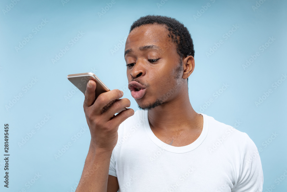 Half-length portrait of young african-american man in white shirt on blue background. Human emotions, facial expression, ad, sales concept. Holding a smartphone, talking or recording a voice message. Half-length portrait of young african-american man in white shirt on blue background. Human emotions, facial expression, ad, sales concept. Holding a smartphone, talking or recording a voice message.
