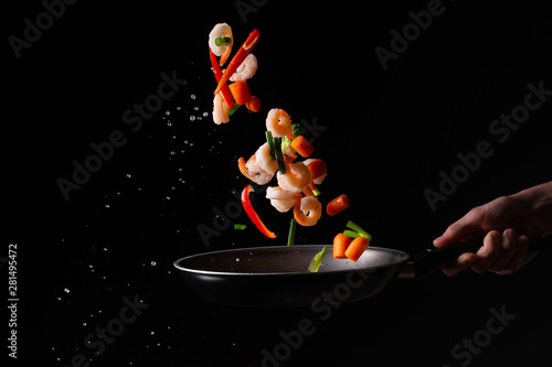Fototapeta Naklejka Na Ścianę i Meble -  close-up, Seafood, chef cooks shrimps with pepper, spiked beans and Brussels sprouts in a frying pan. On a black background for design, menus, restaurants, oriental cuisine, healthy food